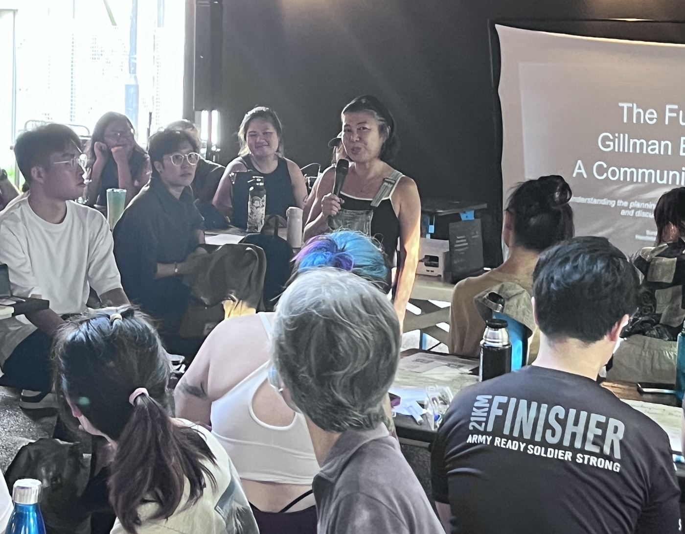A resident speaking into a microphone at the forum, before a slide reading The Future of Gillman Barracks: A Community Dialogue